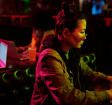 Una persona con gafas trabaja en un portátil en un entorno con poca luz, mientras otra persona usa un portátil al fondo; ambas concentradas en sus tareas, reflejando la cultura de trabajo enfocada de Meta.