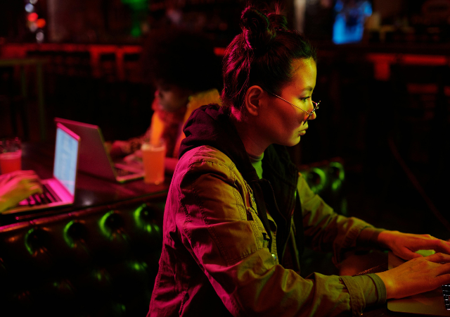 Una persona con gafas trabaja en un portátil en un entorno con poca luz, mientras otra persona usa un portátil al fondo; ambas concentradas en sus tareas, reflejando la cultura de trabajo enfocada de Meta.