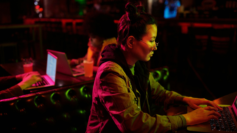 Una persona con gafas trabaja en un portátil en un entorno con poca luz, mientras otra persona usa un portátil al fondo; ambas concentradas en sus tareas, reflejando la cultura de trabajo enfocada de Meta.