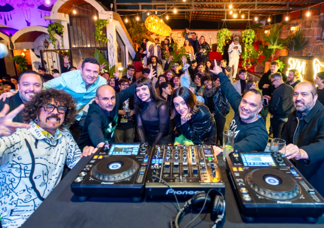 Un grupo de DJ posa para una selfie detrás de los platos en una animada fiesta interior de FIMPro, con una multitud de personas sonriendo y vitoreando al fondo bajo las luces de cadena.