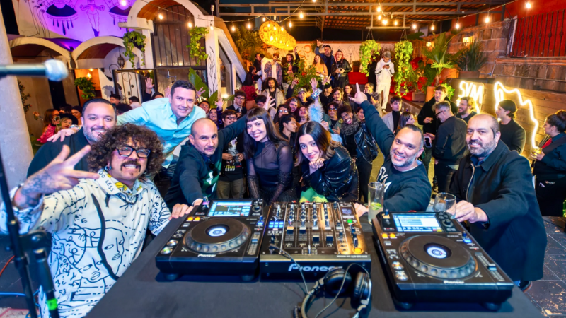 Un grupo de DJ posa para una selfie detrás de los platos en una animada fiesta interior de FIMPro, con una multitud de personas sonriendo y vitoreando al fondo bajo las luces de cadena.