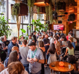 Un gran grupo de personas socializa en un espacio interior de SXSW con paredes de madera, plantas colgantes y luz natural entrando por grandes ventanales.