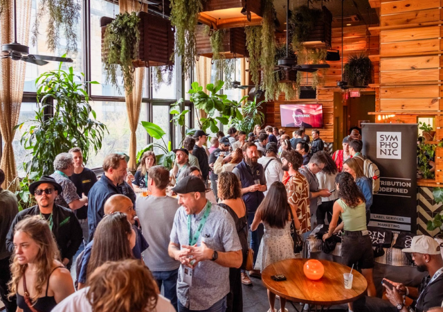 Un gran grupo de personas socializa en un espacio interior de SXSW con paredes de madera, plantas colgantes y luz natural entrando por grandes ventanales.