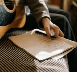 Artista sentado con una guitarra acústica mientras escribe letras en un cuaderno, preparando el lanzamiento de su primera canción.