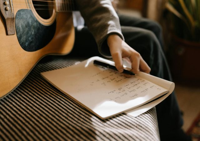 Artista sentado con una guitarra acústica mientras escribe letras en un cuaderno, preparando el lanzamiento de su primera canción.