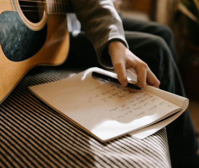 Artista sentado con una guitarra acústica mientras escribe letras en un cuaderno, preparando el lanzamiento de su primera canción.