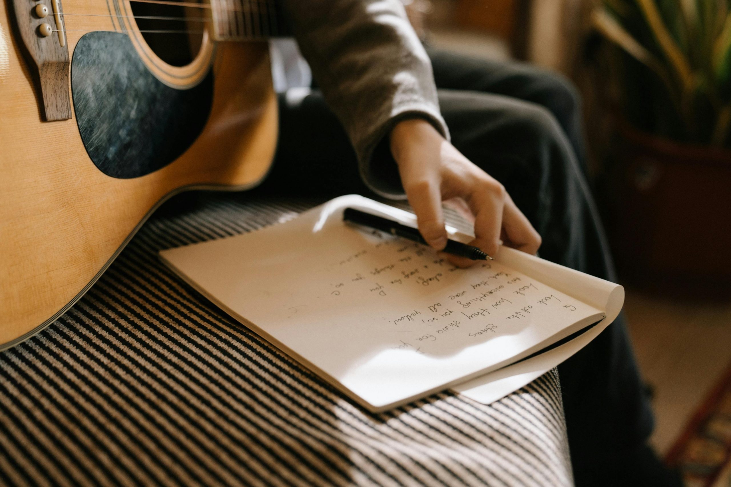 Artista sentado con una guitarra acústica mientras escribe letras en un cuaderno, preparando el lanzamiento de su primera canción.