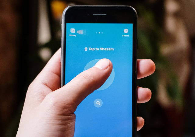Una persona sostiene un smartphone que muestra la app de Shazam con el botón destacado “Tap to Shazam”, listo para el descubrimiento musical.