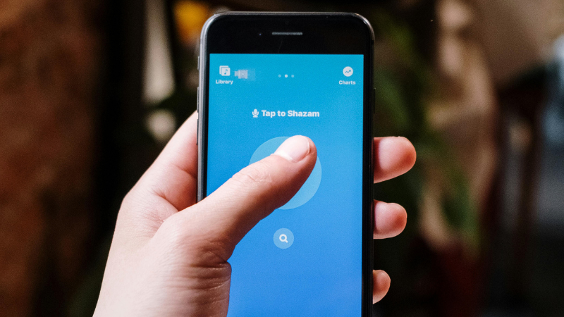 Una persona sostiene un smartphone que muestra la app de Shazam con el botón destacado “Tap to Shazam”, listo para el descubrimiento musical.