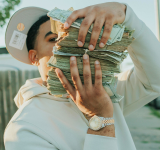 Una persona con sudadera blanca y gorra sostiene varios fajos de dinero en efectivo, cubriendo parcialmente su rostro, mientras lleva un reloj al aire libre, posiblemente celebrando una gran ganancia en music publishing.
