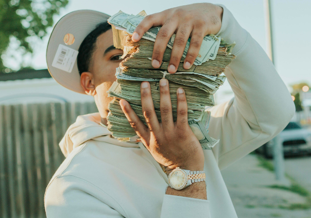 Una persona con sudadera blanca y gorra sostiene varios fajos de dinero en efectivo, cubriendo parcialmente su rostro, mientras lleva un reloj al aire libre, posiblemente celebrando una gran ganancia en music publishing.