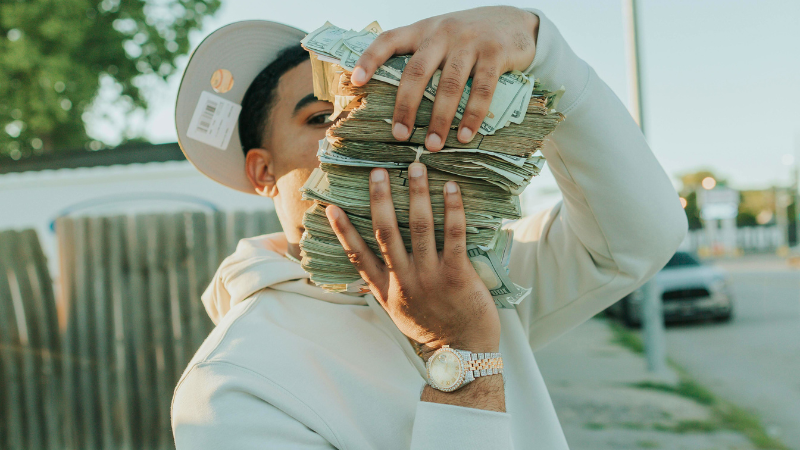 Una persona con sudadera blanca y gorra sostiene varios fajos de dinero en efectivo, cubriendo parcialmente su rostro, mientras lleva un reloj al aire libre, posiblemente celebrando una gran ganancia en music publishing.