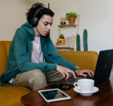 Una persona con audífonos está sentada en un sofá amarillo, usando una laptop para concentrarse en el pitching a playlists, con una tablet y una taza cerca. Plantas y estantes decoran el fondo