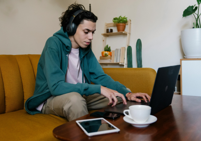 Una persona con audífonos está sentada en un sofá amarillo, usando una laptop para concentrarse en el pitching a playlists, con una tablet y una taza cerca. Plantas y estantes decoran el fondo