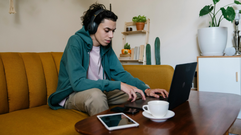 Una persona con audífonos está sentada en un sofá amarillo, usando una laptop para concentrarse en el pitching a playlists, con una tablet y una taza cerca. Plantas y estantes decoran el fondo
