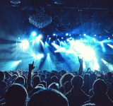 Una gran multitud en un concierto durante una gira, con luces azules en el escenario, manos levantadas y siluetas de personas mirando hacia el escenario iluminado.