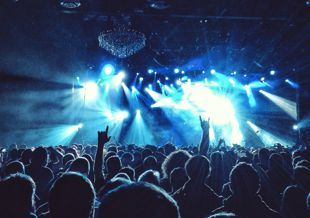 Una gran multitud en un concierto durante una gira, con luces azules en el escenario, manos levantadas y siluetas de personas mirando hacia el escenario iluminado.