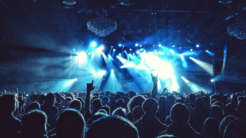Una gran multitud en un concierto durante una gira, con luces azules en el escenario, manos levantadas y siluetas de personas mirando hacia el escenario iluminado.