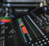 Un primer plano de una consola de mezcla de audio mejorada con IA, con múltiples deslizadores, perillas, botones de colores y una pequeña pantalla. Botellas de agua y otros equipos se ven en el fondo.