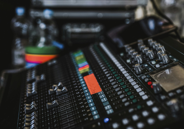 Un primer plano de una consola de mezcla de audio mejorada con IA, con múltiples deslizadores, perillas, botones de colores y una pequeña pantalla. Botellas de agua y otros equipos se ven en el fondo.