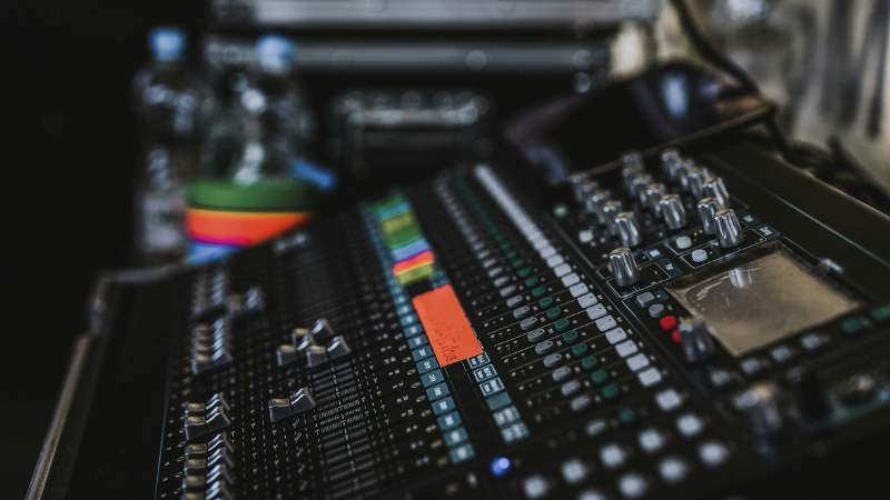 Un primer plano de una consola de mezcla de audio mejorada con IA, con múltiples deslizadores, perillas, botones de colores y una pequeña pantalla. Botellas de agua y otros equipos se ven en el fondo.