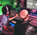 Dos personas trabajan en una consola de mezcla en un estudio de grabación, ajustando niveles de audio mientras observan analíticas de sonido en una gran pantalla de computadora.