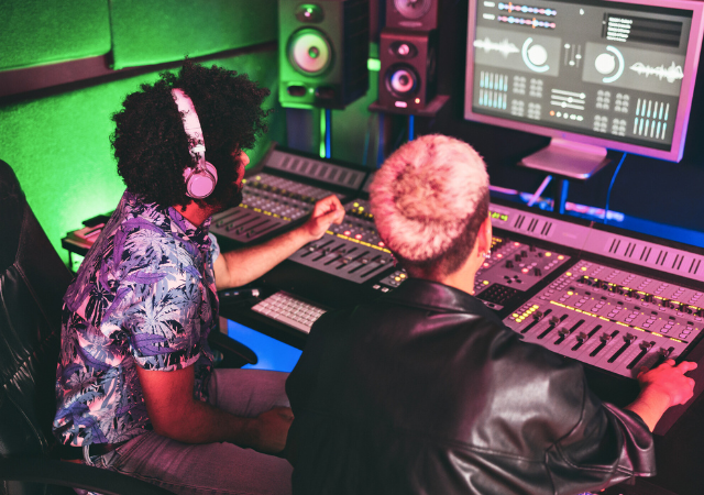 Dos personas trabajan en una consola de mezcla en un estudio de grabación, ajustando niveles de audio mientras observan analíticas de sonido en una gran pantalla de computadora.