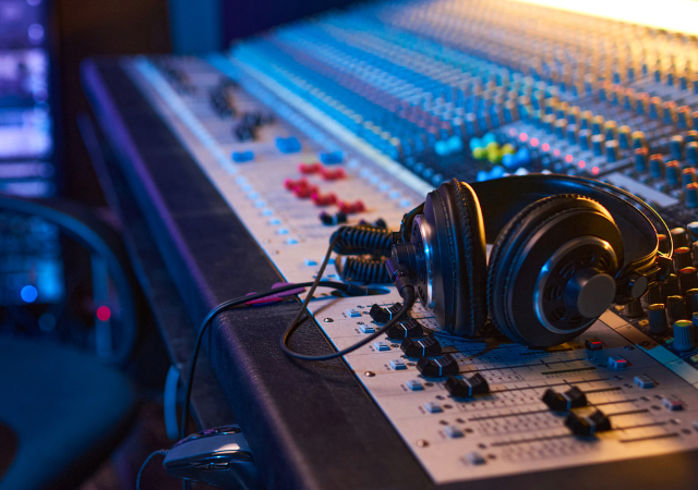 Unos audífonos descansan sobre una gran consola de mezcla de audio con numerosos botones, perillas y faders en un estudio de grabación, donde ingenieros de sonido gestionan datos de audio para lograr grabaciones perfectas.