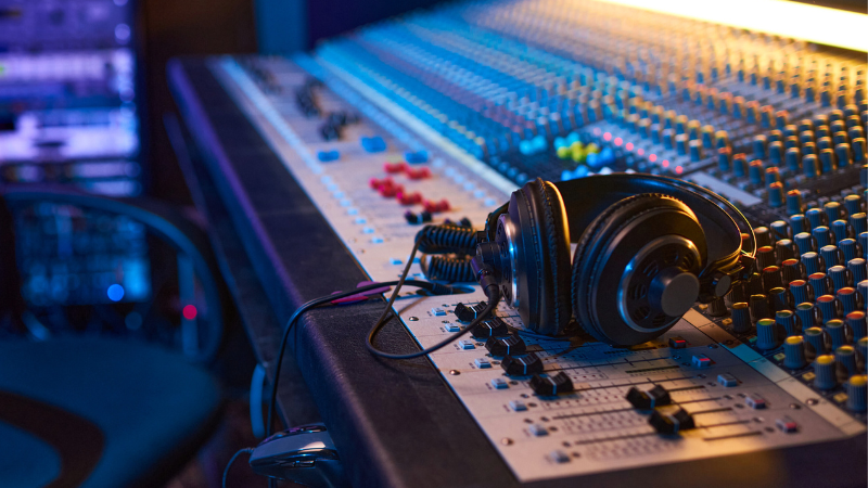 Unos audífonos descansan sobre una gran consola de mezcla de audio con numerosos botones, perillas y faders en un estudio de grabación, donde ingenieros de sonido gestionan datos de audio para lograr grabaciones perfectas.