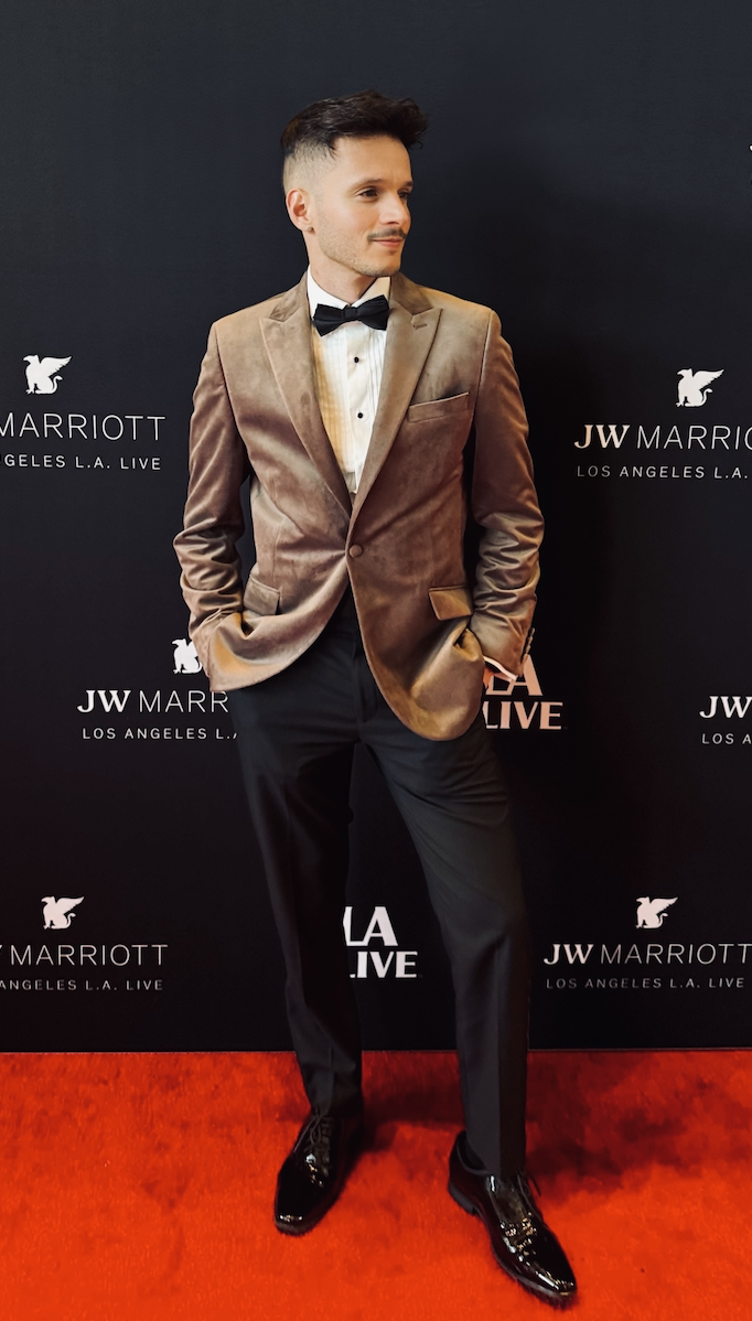 Un hombre con un blazer de terciopelo color beige, camisa blanca, corbatín negro y pantalón negro está en una alfombra roja frente a un fondo de JW Marriott L.A. Live.