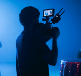 Una persona opera una cámara de video en un set con poca iluminación y luces azules, capturando contenido para YouTube, mientras varias personas aparecen desenfocadas en el fondo.