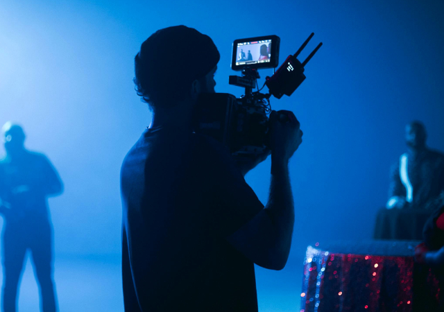 Una persona opera una cámara de video en un set con poca iluminación y luces azules, capturando contenido para YouTube, mientras varias personas aparecen desenfocadas en el fondo.