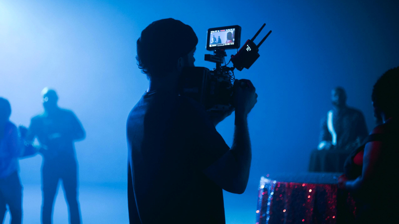 Una persona opera una cámara de video en un set con poca iluminación y luces azules, capturando contenido para YouTube, mientras varias personas aparecen desenfocadas en el fondo.