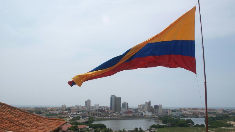 Una gran bandera colombiana ondea sobre el paisaje urbano, donde edificios modernos e históricos se alzan bajo un cielo nublado: una vista inspiradora, especialmente durante Circulart, cuando la creatividad llena el ambiente.