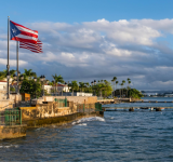 La bandera de Puerto Rico ondea en un paseo marítimo rodeado de palmeras y edificios, mientras el mar tranquilo y el cielo crean el escenario para celebraciones como Premios Tu Música Urbano.