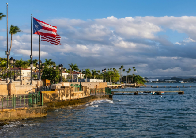 La bandera de Puerto Rico ondea en un paseo marítimo rodeado de palmeras y edificios, mientras el mar tranquilo y el cielo crean el escenario para celebraciones como Premios Tu Música Urbano.