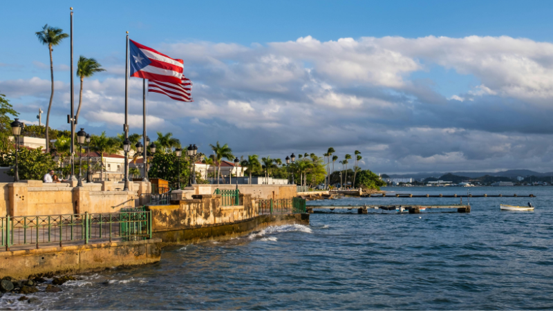 La bandera de Puerto Rico ondea en un paseo marítimo rodeado de palmeras y edificios, mientras el mar tranquilo y el cielo crean el escenario para celebraciones como Premios Tu Música Urbano.