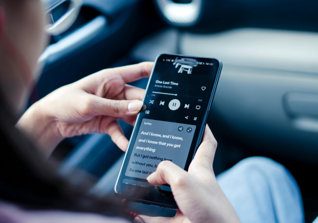 Persona sosteniendo un smartphone dentro de un carro, reproduciendo música y viendo letras en Spotify mostradas en la pantalla.