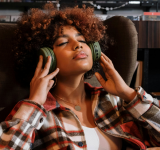 Una persona con cabello rizado está sentada en una silla, con los ojos cerrados, usando grandes audífonos verdes y una camisa de cuadros, aparentando estar relajada mientras escucha música en esta escena editorial.