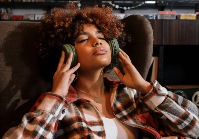 Una persona con cabello rizado está sentada en una silla, con los ojos cerrados, usando grandes audífonos verdes y una camisa de cuadros, aparentando estar relajada mientras escucha música en esta escena editorial.