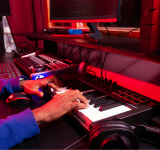 Una persona toca un teclado digital en un estudio de producción musical, creando nuevos tracks con samples en una computadora, equipo de sonido y audífonos sobre un escritorio iluminado con luces rojas.