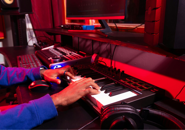 Una persona toca un teclado digital en un estudio de producción musical, creando nuevos tracks con samples en una computadora, equipo de sonido y audífonos sobre un escritorio iluminado con luces rojas.