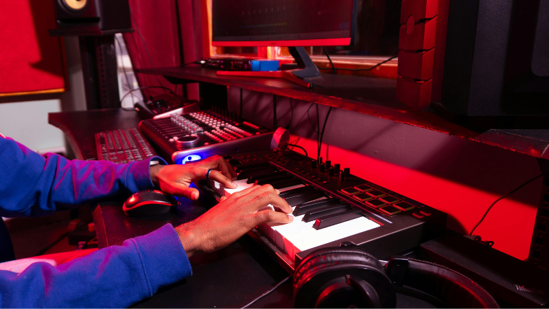 Una persona toca un teclado digital en un estudio de producción musical, creando nuevos tracks con samples en una computadora, equipo de sonido y audífonos sobre un escritorio iluminado con luces rojas.